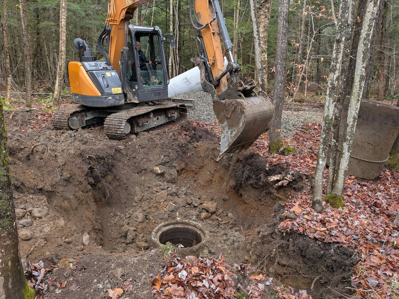 Excavatrice creuse fosse septique en forêt, regard circulaire visible